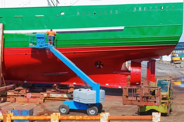 Worker on an elevated platform painting a vessel hull in dry dock